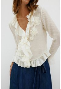 Weißes, strukturiertes Blusenshirt mit Rüschenausschnitt und floralen Akzenten, ausgestattet mit einem geschichteten Saum. Kombination mit einem dunklen Denim-Rock.