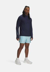 T-shirt de sport bleu marine à manches longues avec un demi-zip, associé à un short bleu clair et des baskets blanches. Caractérisé par un design élégant et fonctionnel.