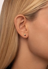 Gold- und schwarze Ohrstecker mit konischer Form oben und flachem schwarzen Fuß, der mit einem kleinen klaren Akzent verziert ist. Am Ohr eines Models getragen.