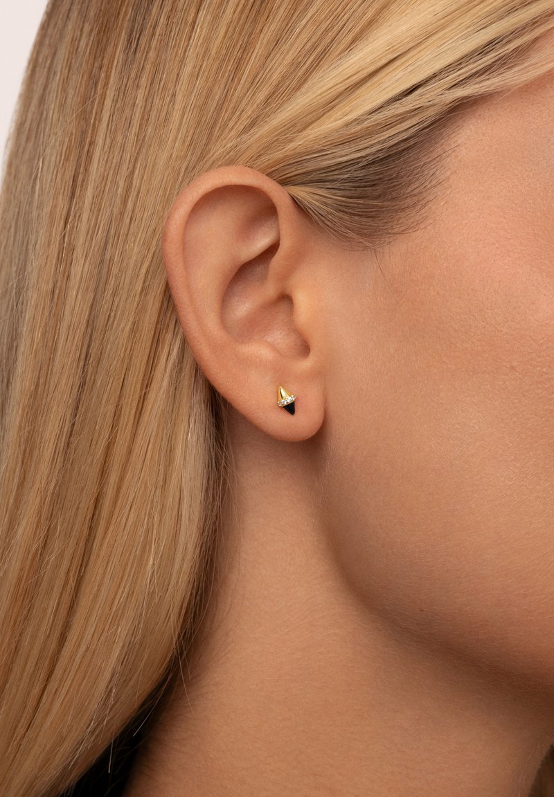 Gold- und schwarze Ohrstecker mit konischer Form oben und flachem schwarzen Fuß, der mit einem kleinen klaren Akzent verziert ist. Am Ohr eines Models getragen.