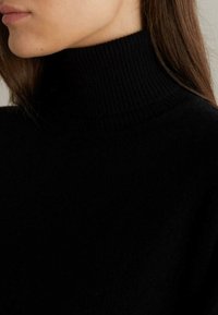 Svart polokrage tröja i mjukt, texturerat tyg med ribbad krage. Designen är figurnära och framhäver en ren, enkel silhuett.