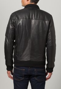 Man in een zwart leren jack met geribbelde manchetten en tailleband, gecombineerd met donkerblauwe jeans, gezien vanaf de achterkant.