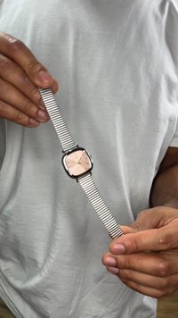 Srebrna zapestna ura s kvadratnim številčnico iz rožnatega zlata, kovinskim mrežastim paščkom in minimalističnim dizajnom. Pašček ima teksturiran vzorec povezanih členov.