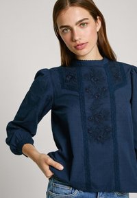 Blusa blu navy con ricamo floreale sulla parte frontale, colletto alto, maniche a sbuffo e dettagli in pizzo lungo le cuciture.