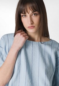 Top texturizado de color azul claro con rayas verticales en blanco y azul. Presenta un escote ancho y un ajuste relajado, exhibiendo detalles de tejido entrelazado.