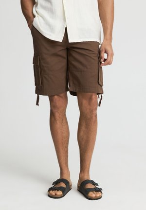 Uomo che indossa pantaloncini cargo marroni, camicia bianca a maniche corte e sandali neri, in piedi davanti a uno sfondo semplice.