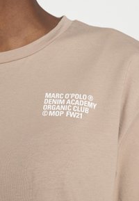 Béžové tričko s kulatým výstřihem, na kterém je bíle tištěný text: "MARC O'POLO® DENIM ACADEMY ORGANIC CLUB © MOP FW21." Měkká bavlněná textura.