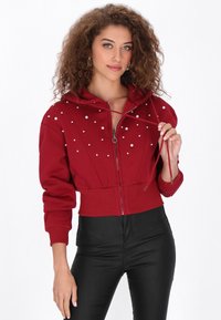 Sudadera corta con cremallera en rojo y capucha, con tachuelas plateadas a lo largo del frente. Combinada con pantalones ajustados negros que muestran un diseño corto.