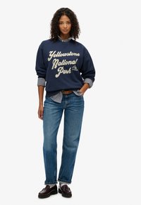 Sudadera azul marino con el texto "Parque Nacional Yellowstone" en crema, superpuesta sobre una camisa de cuello a rayas azul y blanca, combinada con jeans azules.