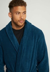 Mörkblå fleece badrock med sjalkrage och knytmidja, med mjuk textur och långa ärmar, bärs över ett svart plagg.
