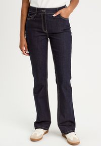 Dark blue denim flared jeans with contrasting yellow stitching, five-pocket design, and a button closure; paired with light sneakers.