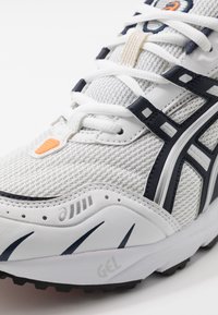 Vit sportskor med mesh-övre, marinblå accenter och orange logotyp. Har vadderad tunga, snören och strukturerad sula. Synlig GEL-märkning.