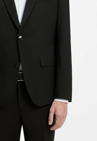 Veste de costume noire sur mesure à deux boutons sur une chemise blanche, pantalon noir assorti et ceinture visible, montrée de la poitrine à la main.