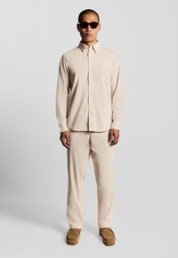 Beige langærmet skjorte med knapper, matchende bukser og brune slip-on sko. Modellen bærer mørke solbriller; glat stof, afslappet pasform.