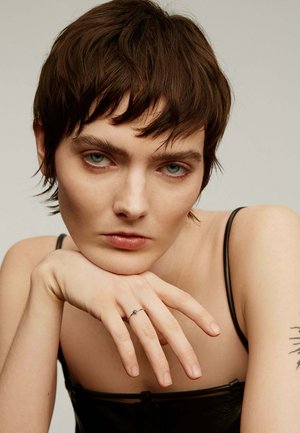 Anillo de plata con una piedra preciosa azul, llevado en un dedo que descansa sobre un hombro desnudo. La figura tiene el cabello corto y marrón y una correa negra en el hombro.