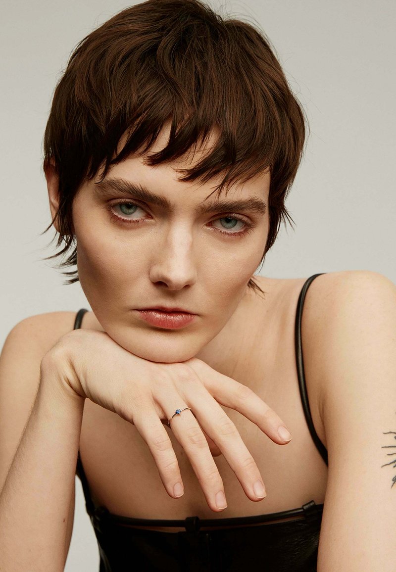 Anillo de plata con una piedra preciosa azul, llevado en un dedo que descansa sobre un hombro desnudo. La figura tiene el cabello corto y marrón y una correa negra en el hombro.