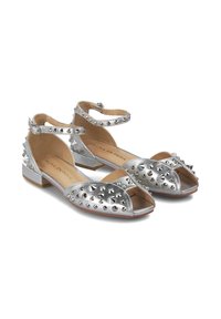 Zapatos planos abiertos de punta plateados con acentos de tachuelas y correas en el tobillo. Presentan un acabado metálico, tacón plano y un diseño abierto para mayor transpirabilidad.