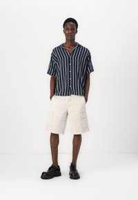 Chemise à rayures navy à manches courtes avec un bouton sur le devant, associée à un short cargo beige doté de poches latérales et à des chaussures noires à lacets.