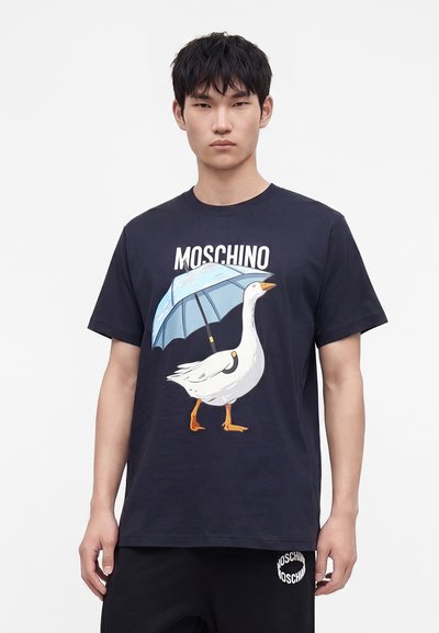 Νεαρός που φοράει μαύρο t-shirt Moschino με γραφικό λευκού χήνα που κρατά μπλε ομπρέλα και μαύρο παντελόνι που ταιριάζει.