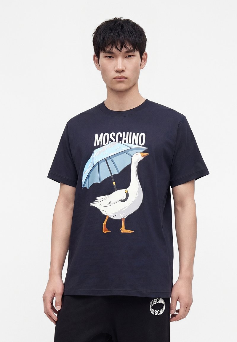 Νεαρός που φοράει μαύρο t-shirt Moschino με γραφικό λευκού χήνα που κρατά μπλε ομπρέλα και μαύρο παντελόνι που ταιριάζει.