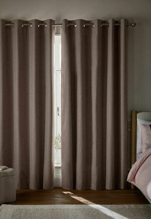 Tende oscuranti marroni appese a una barra di metallo coprono una finestra lasciando entrare un fascio stretto di luce sul pavimento in legno vicino a un letto e un pouf.