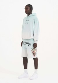 Lyseblå gradient hættetrøje med kængurulomme; koordinerende shorts med grafisk design og sort skrift; parret med hvide sneakers.