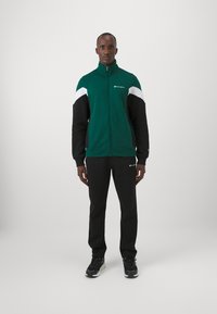Giacca felpata verde con zip, decorata con dettagli in bianco e nero, abbinata a pantaloni sportivi neri. Tessuto spesso con vestibilità comoda, completa di dettagli con logo.