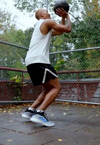Pantaloni sportivi bianco e nero, canotta bianca senza maniche e scarpe da ginnastica con dettagli blu. Palla tenuta sopra la testa; campo all'aperto con rete a maglie.