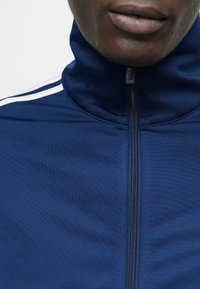 Μπλε σκούρο μπουφάν με φερμουάρ, με ψηλό γιακά, υφή υφάσματος και λευκές ρίγες κατά μήκος των ώμων. Κοντινή λήψη που τονίζει τις λεπτομέρειες.