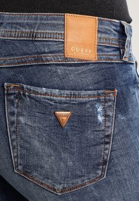 Vaqueros de mezclilla en azul oscuro con ligero desgaste, con una etiqueta de cuero marrón grabada con "GUESS" y un logo triangular en el bolsillo trasero.