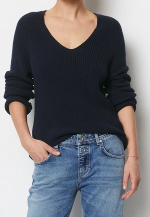 Kobieta ubrana w ciemnogranatowy sweter z dzianiny z dekoltem w kształcie litery V i podwiniętymi rękawami oraz jasnoniebieskie jeansy z guzikiem, z jedną ręką w kieszeni, na jednolitym tle.