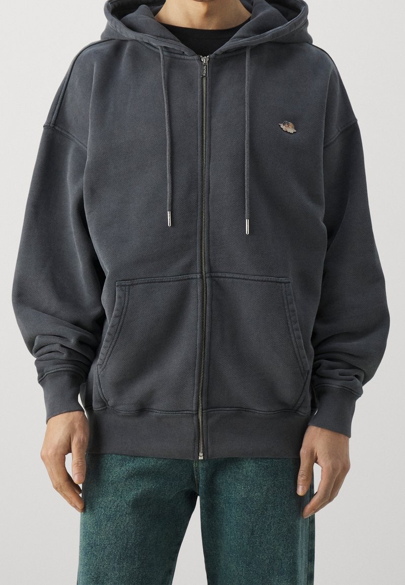 Uomo che indossa un hoodie grigio scuro con zip, con cordini e tasche kangaroo, abbinato a pantaloni verdi con texture.
