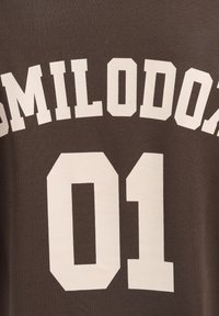 Brun bomulds t-shirt med stor cremefarvet tekst, der står "SMILODON" over nummeret "01." Blød struktur med et afslappet design.