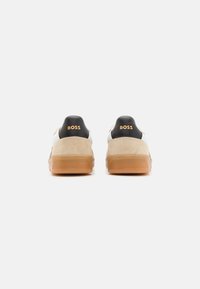Sneaker bianche e beige con dettagli neri sul tallone. Presentano materiali in pelle liscia e suede. Logo "BOSS" in oro sul retro.