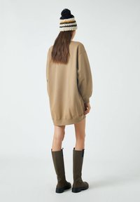 Kobieta odwrócona plecami, ubrana w beżową, oversize'ową sukienkę swetrową, w paskowaną wełnianą czapkę z pomponem oraz oliwkowo-zielone, sięgające do kolan botki na białym tle.