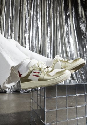 Hvide og guld Tommy Jeans sneakers båret med hvide sokker og frynsede hvide bukser, hvilende på en refleksiv gitterkubus mod en metallisk sølv baggrund.