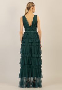 Gr�ünes Tüllkleid mit schichtigen Rüschen, tiefem V-Ausschnitt im Rücken, tailliertem Schnitt und kurzen Trägern. Der Saum reicht bis zum Boden und wird von schwarzen Absatzschuhen ergänzt.
