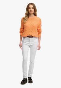 Orange kabelstrikket sweater med lange ærmer, parret med tætsiddende hvide jeans og sorte snørebåndssko. En simpel bælte fremhæver taljen.