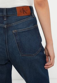 Jeans en denim foncé avec une coupe droite, arborant une patch en cuir marron sur la taille et des accents de couture contrastants.