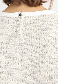 Gros plan de l'encolure arrière d'un pull en maille crème texturée avec un seul bouton noir et des cheveux courts bruns visibles au-dessus.