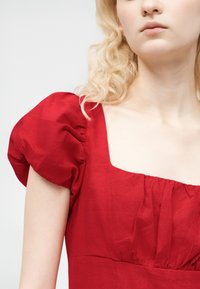 Personne blonde portant une robe rouge à manches courtes bouffantes et décolleté carré, visible des épaules jusqu'au haut de la poitrine sur un fond uni.