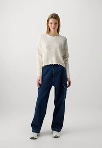 Jeune femme portant un haut blanc texturé à manches longues, un pantalon cargo bleu foncé ample et des baskets blanches, se tenant devant un fond uni.