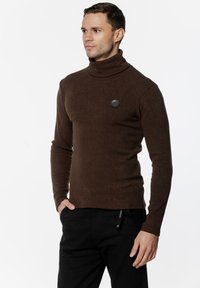 Brązowy sweter z golfem o prążkowanej teksturze, z okrąłym czarnym logo na piersi. Noszony z czarnymi spodniami.