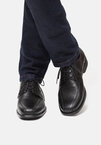 Scarpe da uomo in pelle nera con punta arrotondata e lacci, abbinate a jeans scuri in denim. Le scarpe hanno una superficie liscia e dettagli di cucitura sottili.