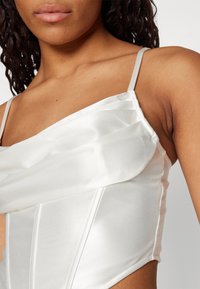 Vit satin-topp med en strukturerad design, som har tunna band och draperade tygdetaljer över bysten och sidorna. Slät textur.