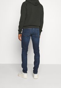 Mężczyzna w ciemnozielonej bluzie z kapturem, w slim fit ciemnoniebieskich dżinsach i białych trampkach Reebok, stojący na tle jednolitego białego tła.