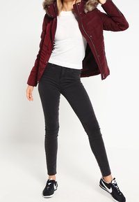 Vinröd puffjacka med huva av fuskpäls, kombinerad med en vit stickad topp och svarta tajta jeans. Svarta sneakers fulländar outfiten.