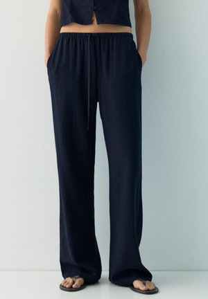 Személy laza fekete húzózsinóros nadrágot és fekete papucsot viselve, egy sima világos falnak támaszkodva, kezét a zsebében tartva áll.