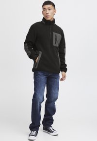 Schwarzer Fleece-Pullover mit grauen Akzenten, zippered Brusttasche und Seitentaschen. Getragen mit blauen Jeans und schwarzen Sneakers.