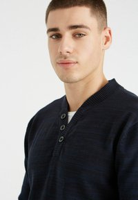 Suéter de punto en color navy con cuello de canalé, tapeta de tres botones y textura sutil. Lleva puesto sobre una camisa blanca, mostrando un primer plano del escote.